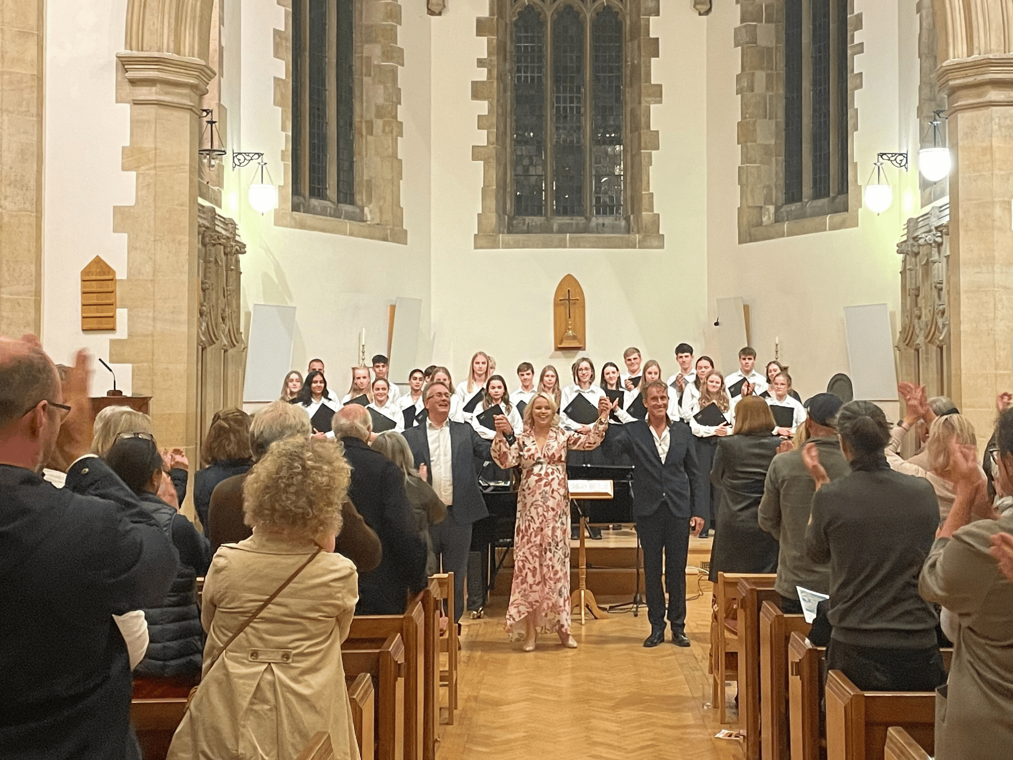 A concert of two singers and a pianist who are bowing and a choir in the background
