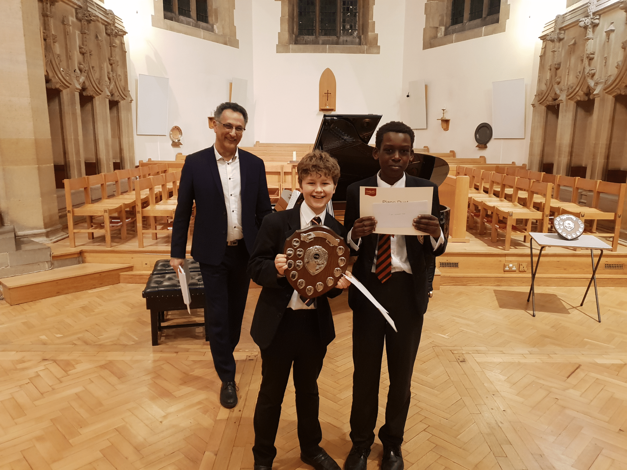 Two students smile with certificates and a shield after winning piano competition