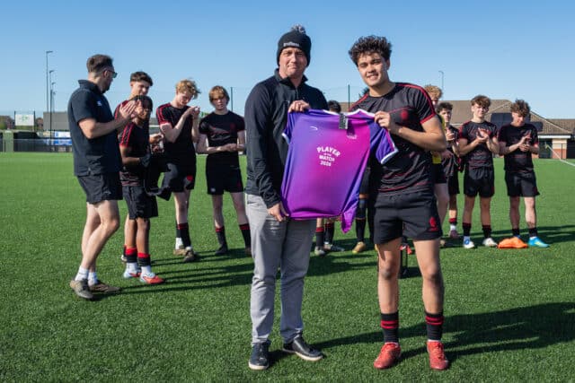 Two people - one in casual clothes with a bobble hat saying Limitless and the other in football kit - holding a football shirt with other boys in football kit applauding him