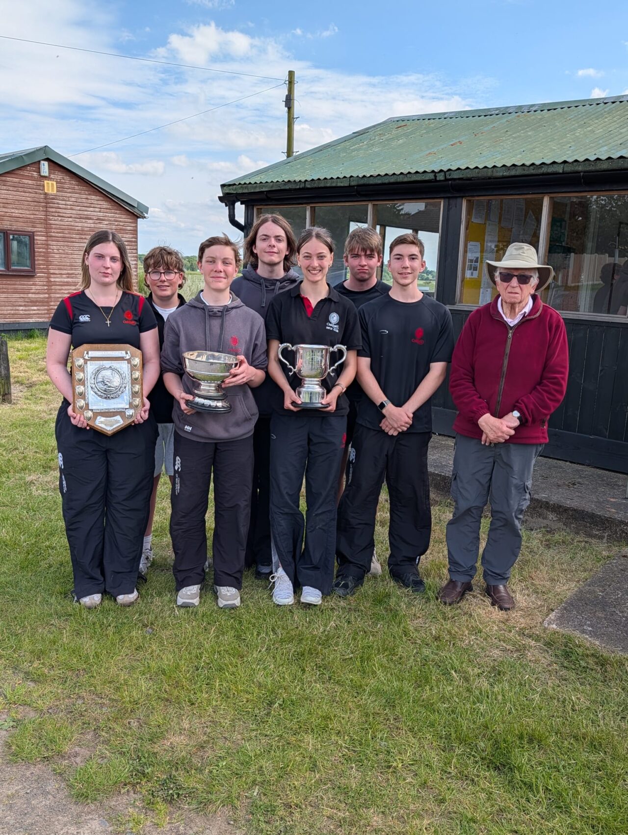 A group of seven students and an older man in a burgundy top and grey hat standing together and holding three cups.