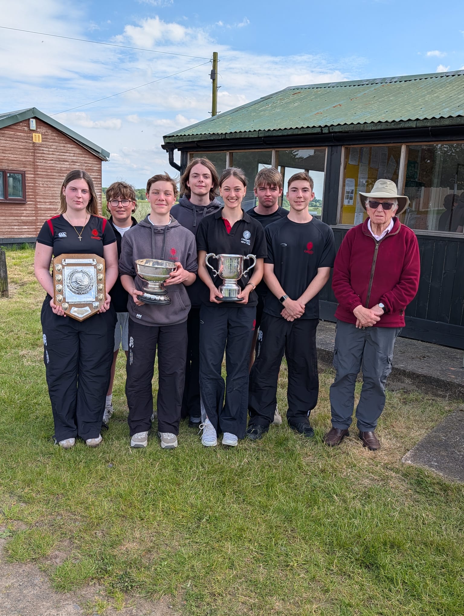 A group of seven students and an older man in a burgundy top and grey hat standing together and holding three cups.