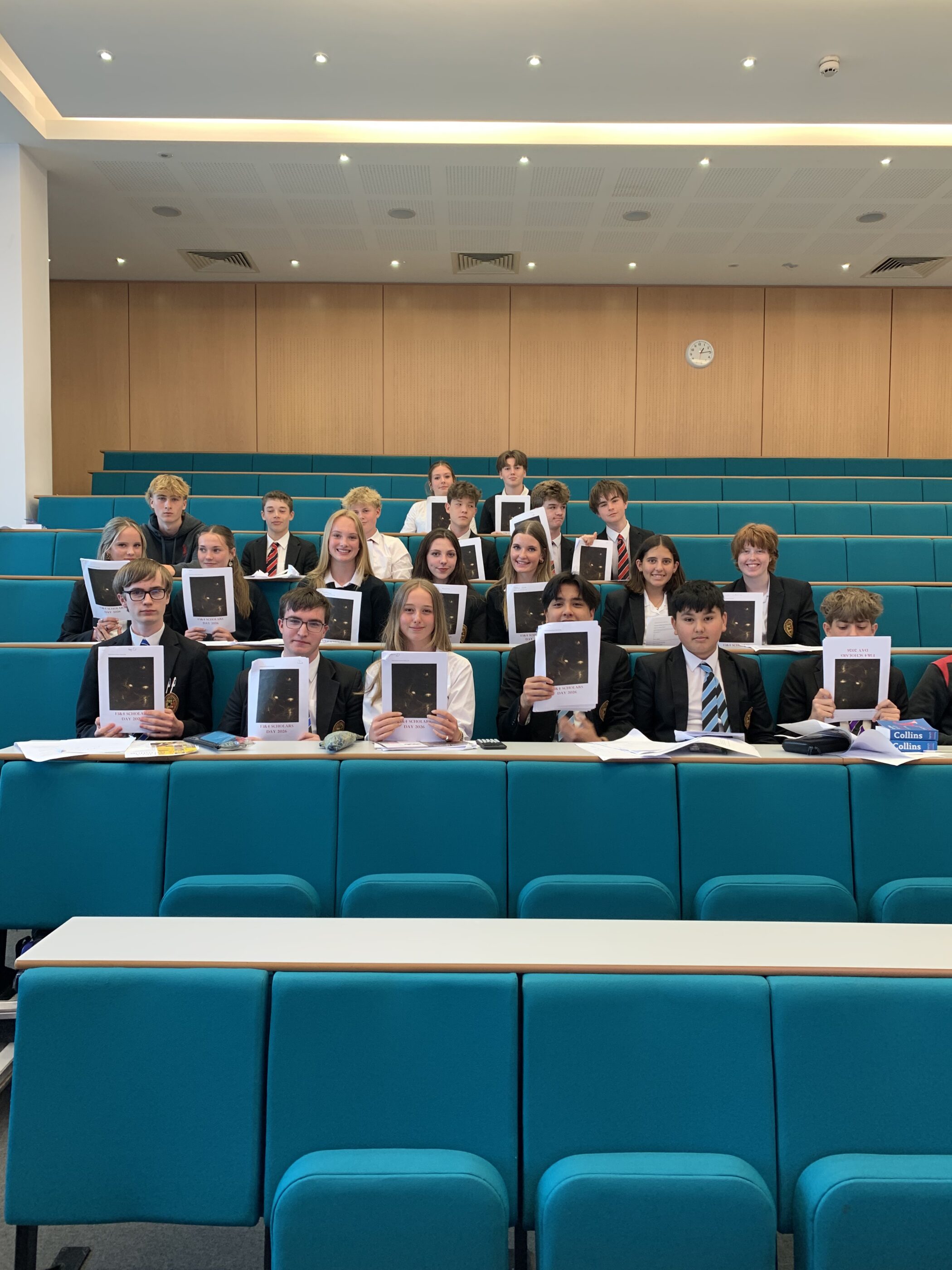 A group of teenagers wearing school uniform sitting in a lecture hall holding up a piece of A4 paper.